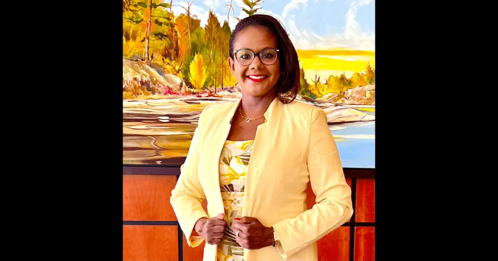 Minerva Ward stands confidently in a yellow jacket with a colorful landscape painting in the background, discussing Saint Lucia Government Crime Inaction.