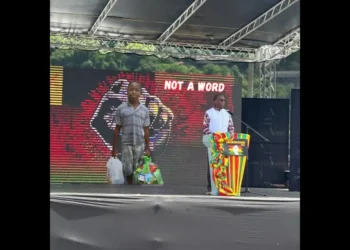 Saint Lucia government silence illustrated as Philip J. Pierre speaks at a podium on Emancipation Day with a backdrop of Young Melchizedek Tixcy holding grocery bags reading Not a Word.
