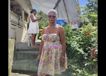 Angela Charlemagne Dies at 73, pictured in a colorful floral dress standing in a lush St Lucia garden