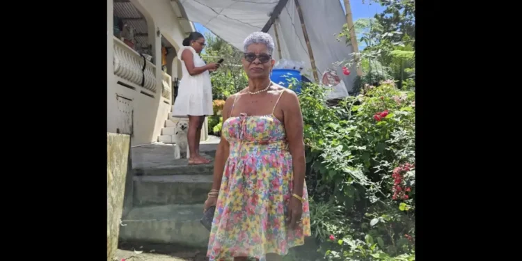 Angela Charlemagne Dies at 73, pictured in a colorful floral dress standing in a lush St Lucia garden