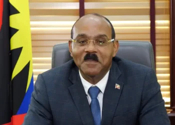 Prime Minister Gaston Browne assures citizens while seated at his desk during a New Year’s address, with national flags behind him in Antigua and Barbuda.