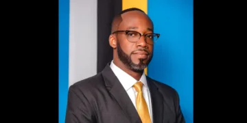 Elisha Norbert wearing a suit and gold tie in a formal portrait against a blue background