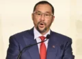 Stuart Young Record article features Stuart Young speaking at a podium in a blue suit, red tie and glasses