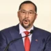Stuart Young Record article features Stuart Young speaking at a podium in a blue suit, red tie and glasses
