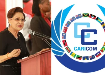 Kamla Persad-Bissessar speaking at a podium alongside the Caricom logo representing the T&T defies Caricom reappointment dispute.