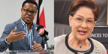 Dr. Amery Browne speaking at a podium beside Prime Minister Kamla Persad-Bissessar in Caricom Barnett row image.