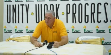 Allen Chastanet seated before a UWP Unity, Work, Progress backdrop as commentary examines UWP internal divisions and party leadership.